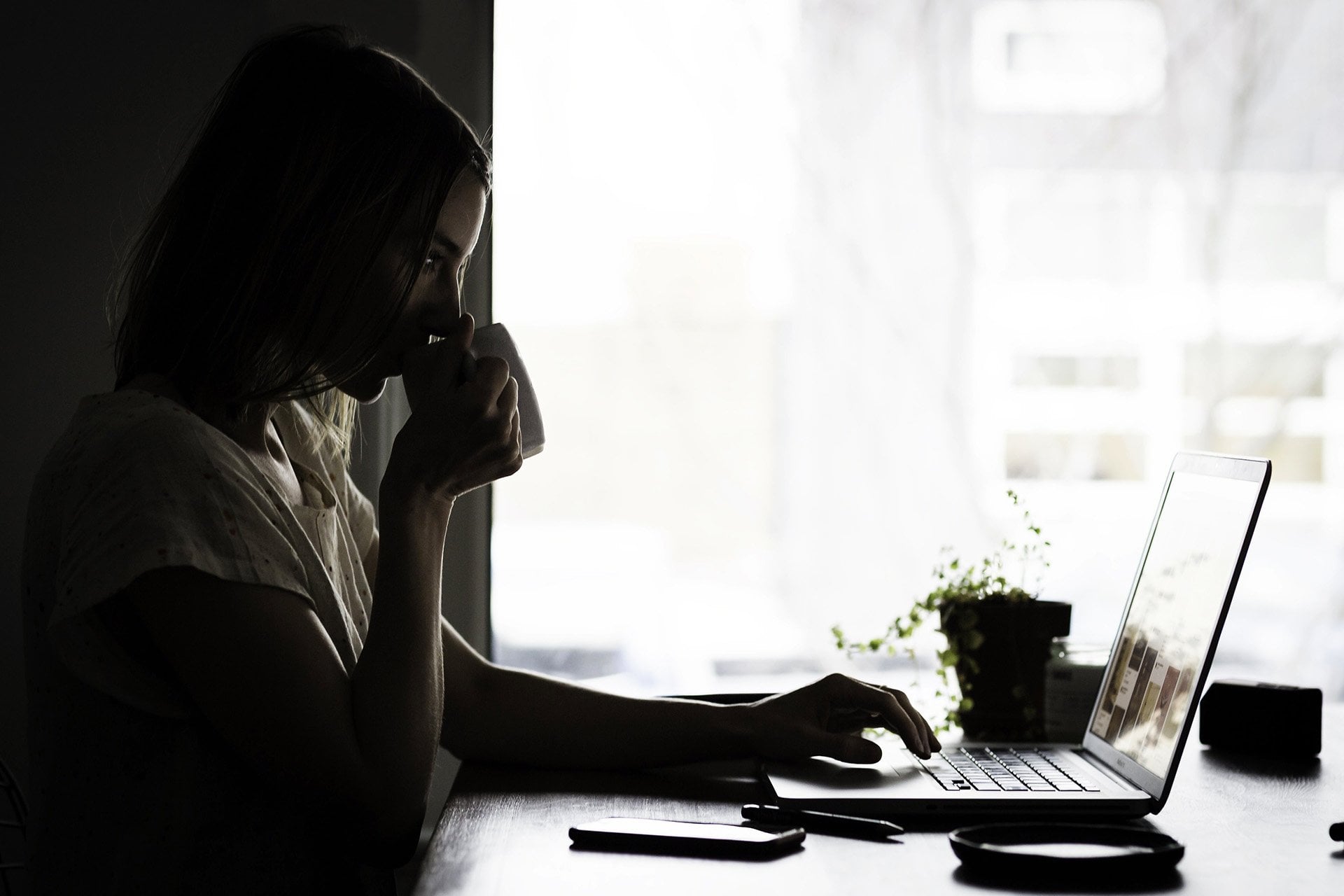 woman drinking coffee looking at a laptop in front of a window